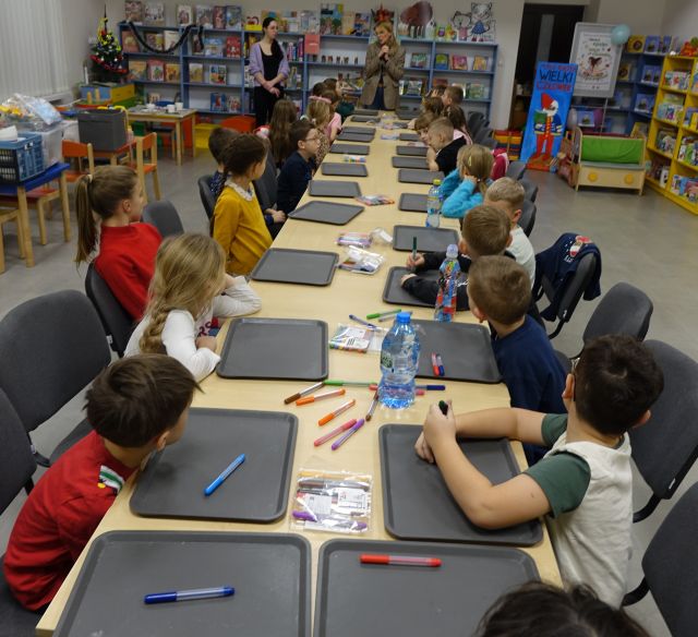 25 dzieci uczestniczących w warsztatach siedzą przy stołach, na których leżą siwe tacki i flamastry
