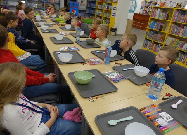 25 dzieci siedzi przy stołąch, na któych leżą siwe tacki i miseczki