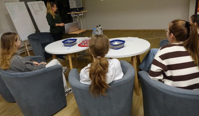 młodzież siedząca na siwych fotelach przy okrągłych białych stołach ogląda prezentację wyświetlaną na ścianie