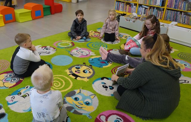 6 dzieci siedzi na kolorowym dywanie, bibliotekarka czyta im książkę
