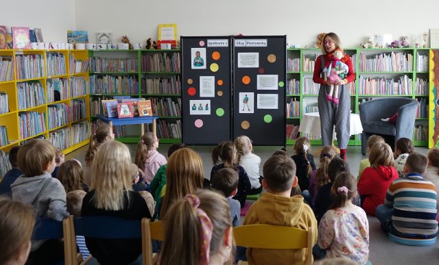 sala biblioteczna wypełniona dziećmi uczestniczącymi w spotkaniu autorskim, przed nimi autorka, w tle regały z książkami