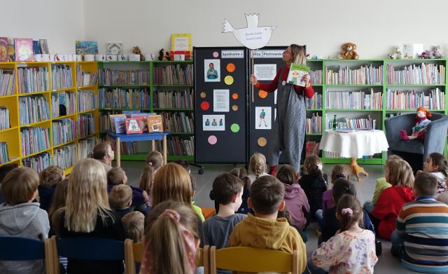 sala biblioteczna wypełniona dziećmi uczestniczącymi w spotkaniu autorskim, przed nimi autorka, w tle regały z książkami