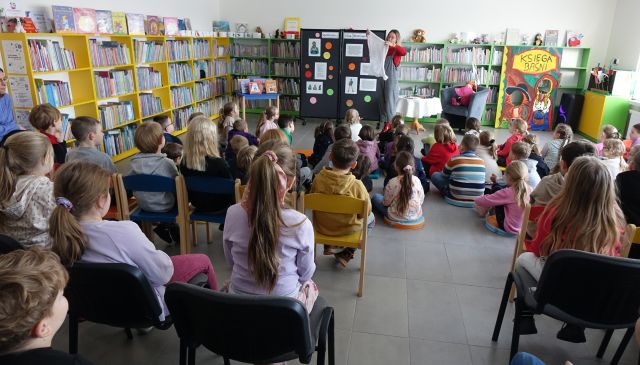 dzieci uczestniczące wspotkaniu autorskim siedzą na krzesłąch, przed nimi autorka, w tle regały z książkami