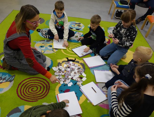 dzieci i autorka siedzą na kolorowym dywanie i wypisują pocztówki