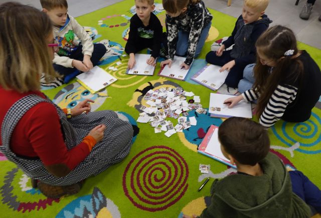 dzieci i autorka siedzą na kolorowym dywanie i wypisują pocztówki