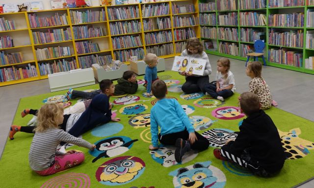 dzieci siedzą na kolorowym dywanie i słuchają czytanej przez bibliotekarkę książki
