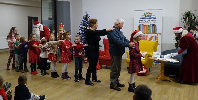 dzieci i dwie starzse osobty stoją jeden za drugim trzymając sie za ramiona, w tle choinka