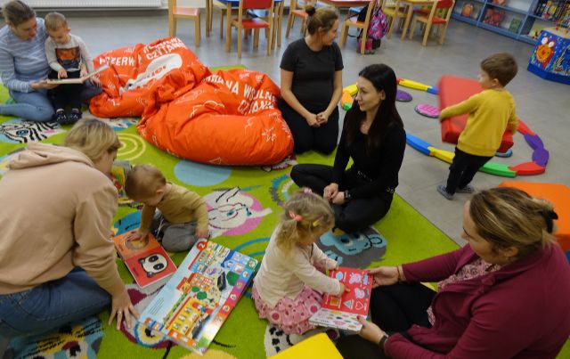 dzieci z mamusiami siedzą na kolorowym dywanie i oglądają książki