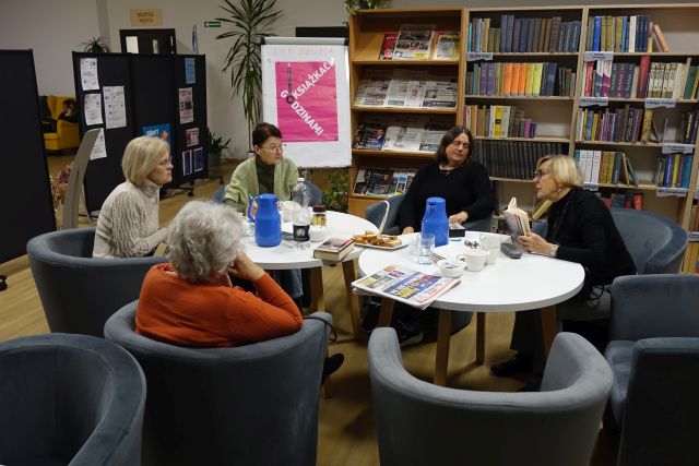 5 kobiet siedzi na siwych fotelach i dyskutuje, w tle regały z książkami