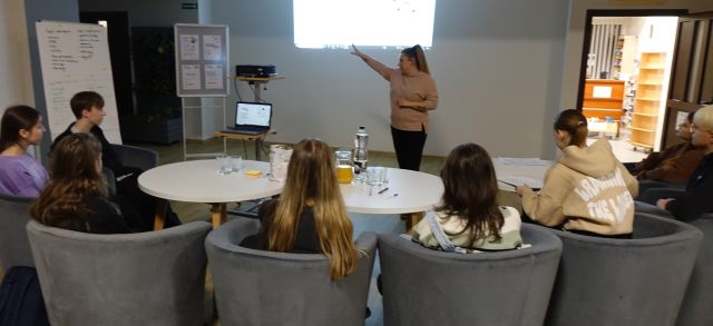 młodzież siedzi na siwych fotelach przy białych okrągłych stołach, bibliotekarka stoi przed nimi, w tle prezentacja na ekranie