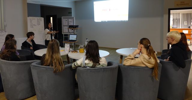 młodzież siedzi na siwych fotelach przy białych okrągłych stołach, bibliotekarka stoi przed nimi, w tle prezentacja na ekranie