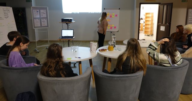 młodzież siedzi na siwych fotelach przy białych okrągłych stołach, bibliotekarka stoi przed nimi, i przegląda kolorowe kartki na flipcharcie