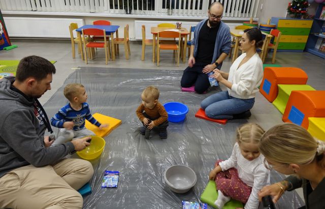 dzieci i rodzice siedzą na podłodze na siwej folii malarskiej, przed nimi leżą miski