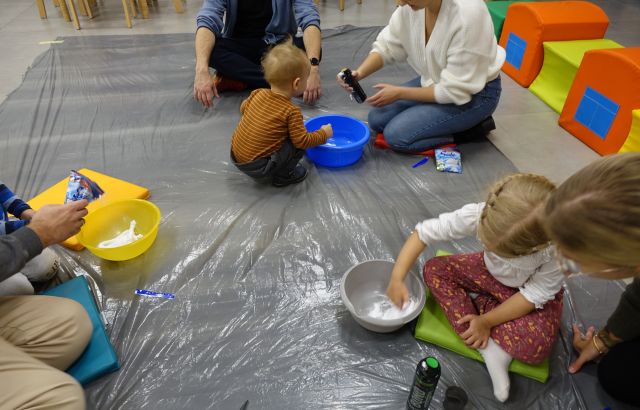 3 małych dzieci, 2 mamy i 2 tatusiów siedzi na podłodze i robią śnieg w miskach z pianki do golenia