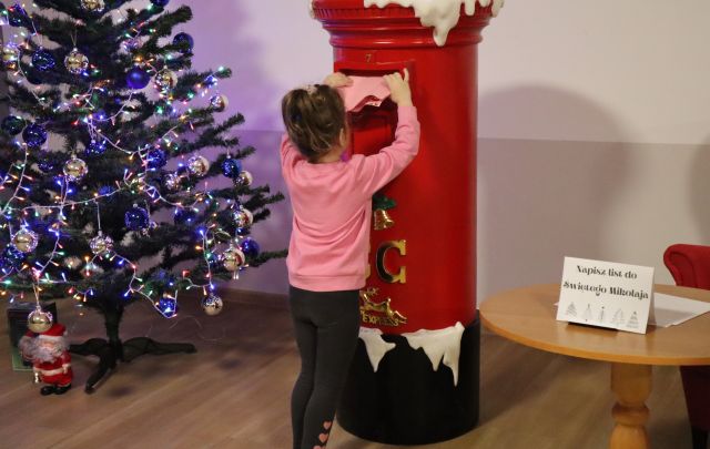 dziewczynka wrzuca list do mikołaja do dużej czerwonej skrzynki, z lewej strony choinka ubrana w lamki i białę i niebieskie bombki