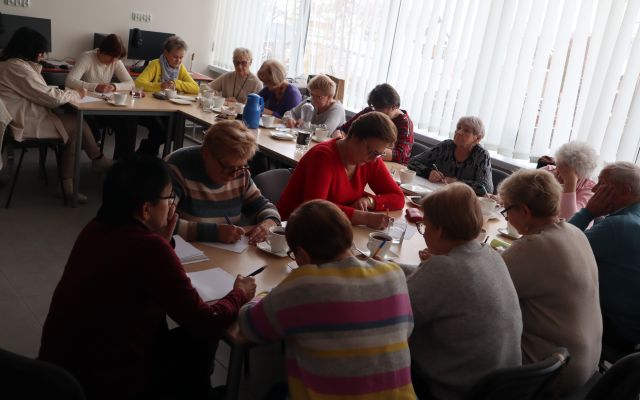 16 starszych osób siedzi przy stołach i pisze