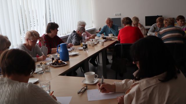 16 starszych osób siedzi przy stołach i pisze