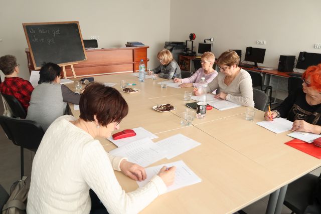 7 seniorek siedzi przy połączonych stołach, na stołach leżą kartki, seniorki piszą