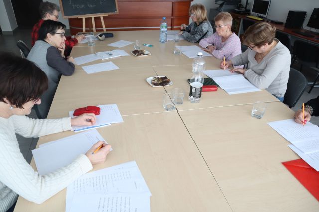 7 seniorek siedzi przy połączonych stołach, na stołach leżą kartki