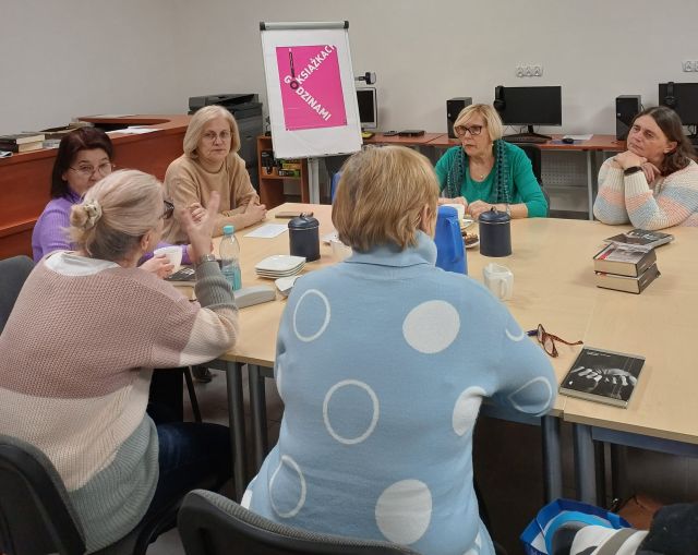 6 kobiet siedzi przy stole i rozmawia, na stole niebieski termos i ciastka na talerzykach, w tle komputery i plakat dkk