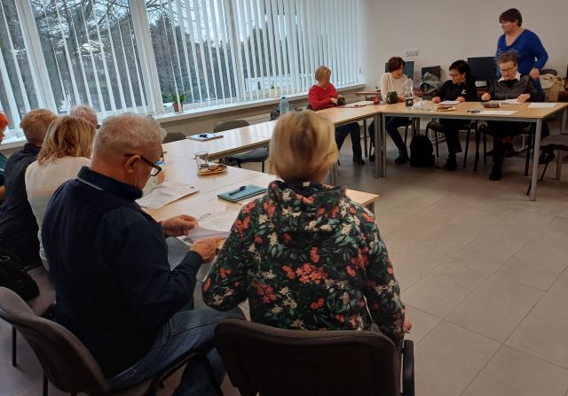 Seniorzy siedzą przy stołach ustawionych w podkowę i rozmawiają