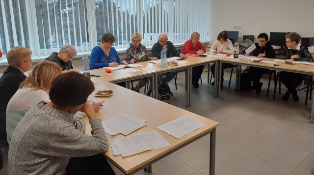 seniorzy siedzą przy stołach ustawionych w podkowę i notują