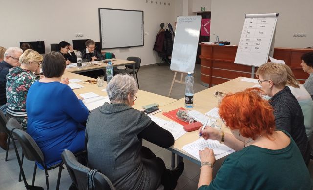 seniorzy siedzą przy stołach ustawionych w podkowę i notują, w tle komputery, flipchart, tablica interaktywna, biurko