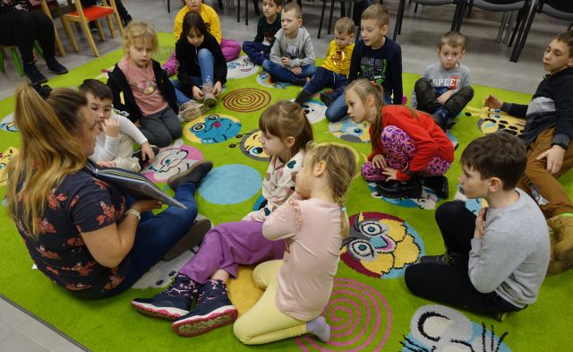 dzieci i bibliotekarka z książką siedzą na kolorowym dywanie