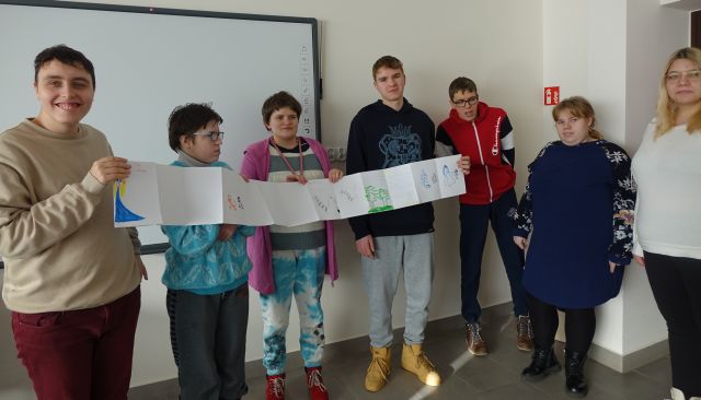 7 osób stoi i w dłoniach trzyma długą książką harmonijkową