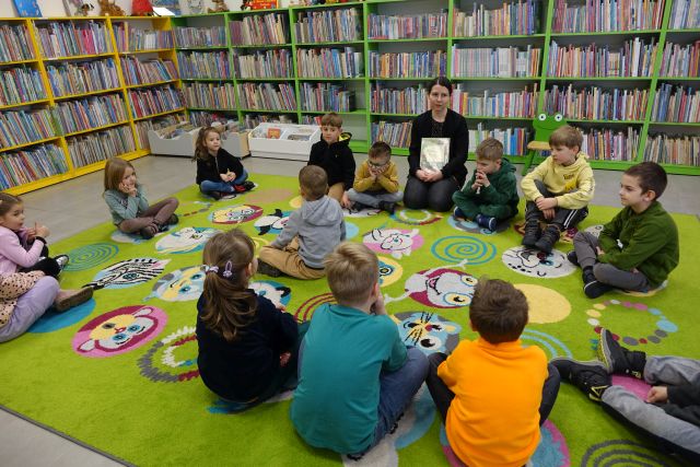 dzieci i bibliotekarka z ksiązką siedzą na kolorowym dywanie, w tle regały z książkami