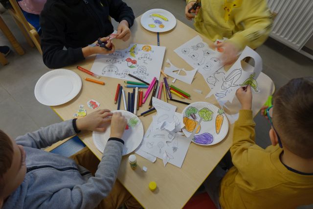 dzieci siedzące przy stolikach malują, kleją i wycinają owoce i warzywa