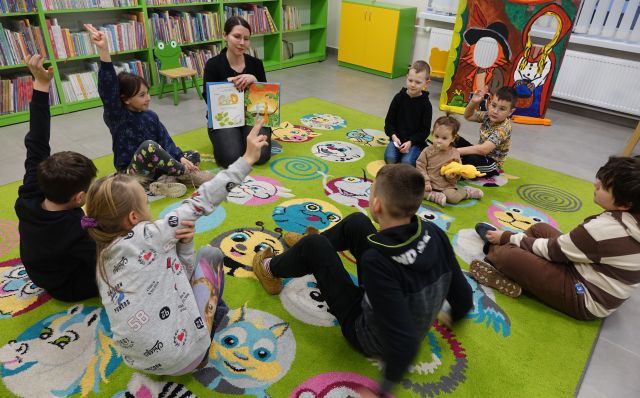 przed bibliotekarką siedzącą na kolorowym dywanie pokazującą książkę siedzą dzieci, w tle regały z książkami