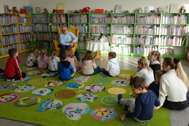 przedszkolaki siedzą na kolorowym dywanie przed starszą kobietą siedzącą na żółtym fotelu, w tle regały z książkami