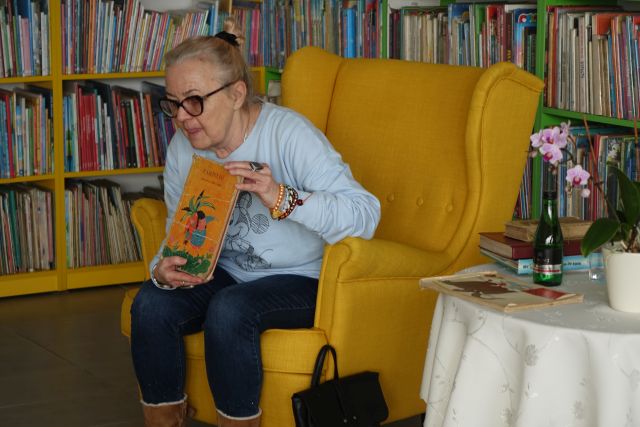 starsza kobieta siedząca na żółtym fotelu pokazuje dzieciom książkę