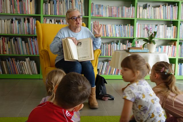 kobieta siedząca na żółtym fotelu pokazuje dzieciom siedzącym na kolorowym dywanie książkę