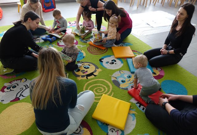 małe dzieci i rodzece siedzą na kolorowym dywanie i oglądają książki