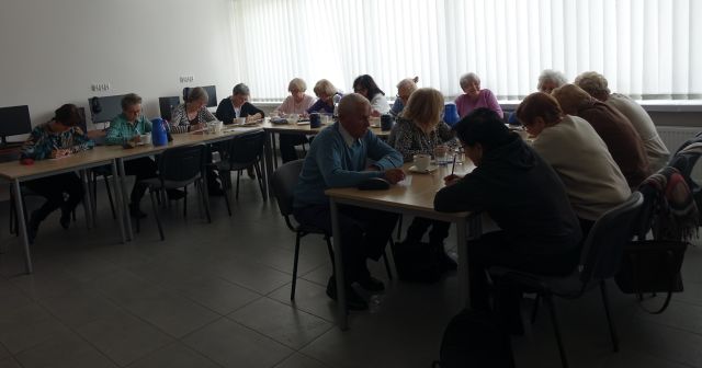 około dwudziestoosobowa grupa seniorów siedzi przy stołach ustawionych w podkowę