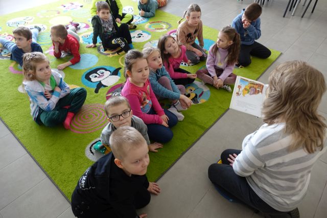 dzieci siedzxą na kolorowym dywanie i słuchają czytającej bibliotekarki