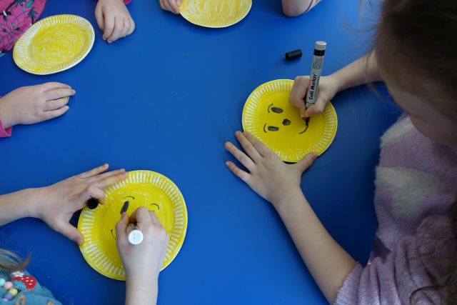 dziecięce rączki malujące oczu, usta i nos na pomalowanych na żółto papierowych talerzykach