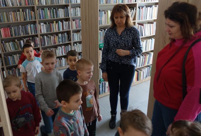 przedszkolaki spacerują z dyrektorką biblioteki pomiędzy regałami z książkami