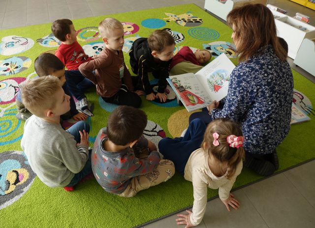 dzieci siedzą na kolorowym dywanie i słuchają czytanej przez dyrektorkę biblioteki książki