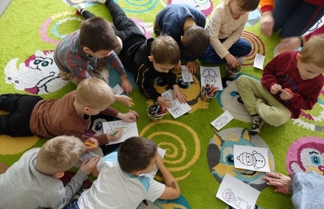dzieci siedzą i leżą na kolorowym dywanie i kolorują obrazki