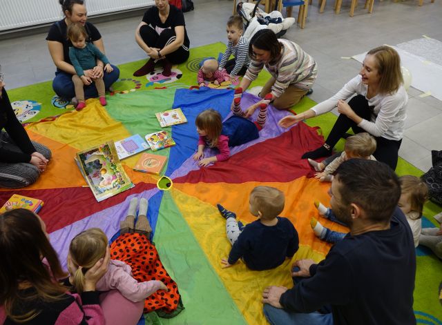 małę dzieci i rodzice siedzą na kolorowej chuście animacyjnej i oglądają kisążki dla dzieci