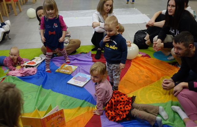 małę dzieci i rodzice siedzą na kolorowej chuście animacyjnej i oglądają kisążki dla dzieci