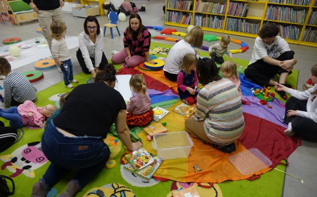 małe dzieci z rodzicami bawią się siedząc na kolorowej chuście animacyjnej
