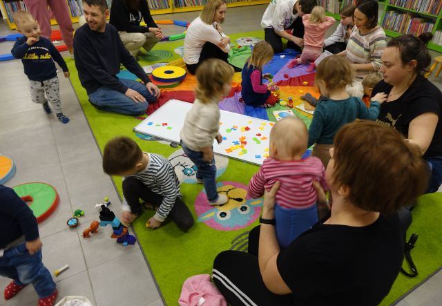 małe dzieci z rodzicami bawią się siedząc na kolorowej chuście animacyjnej