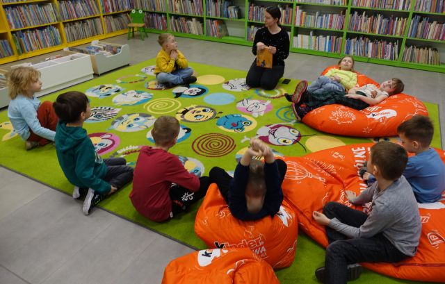 dzieci i bibliotekarka siedzą na kolorowym dywanie, w tle regały z książkami