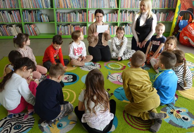 przedszkolaki, bibliotekarka i przedszkolanka siedzą na kolorowtym dywanie, w tle regały z książkami