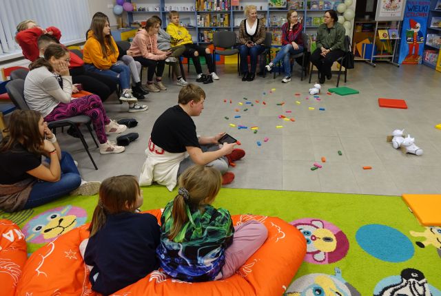 młodzież i dzieci siedzą na krzesłach i podłodze i sterują przy użyciu tabletów robotami Photon