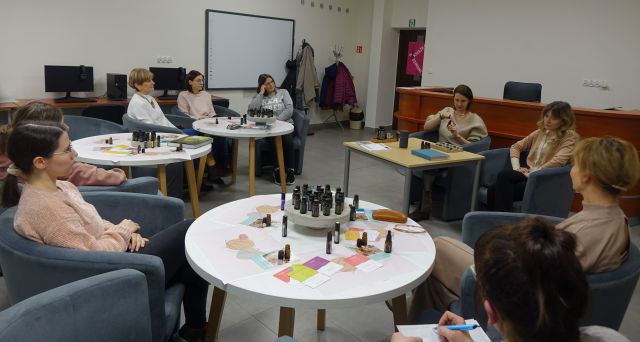 uczestnicy warsztatów olejkowych siedzą na siwych fotelach przy białych okrągłych stołach z olejkami